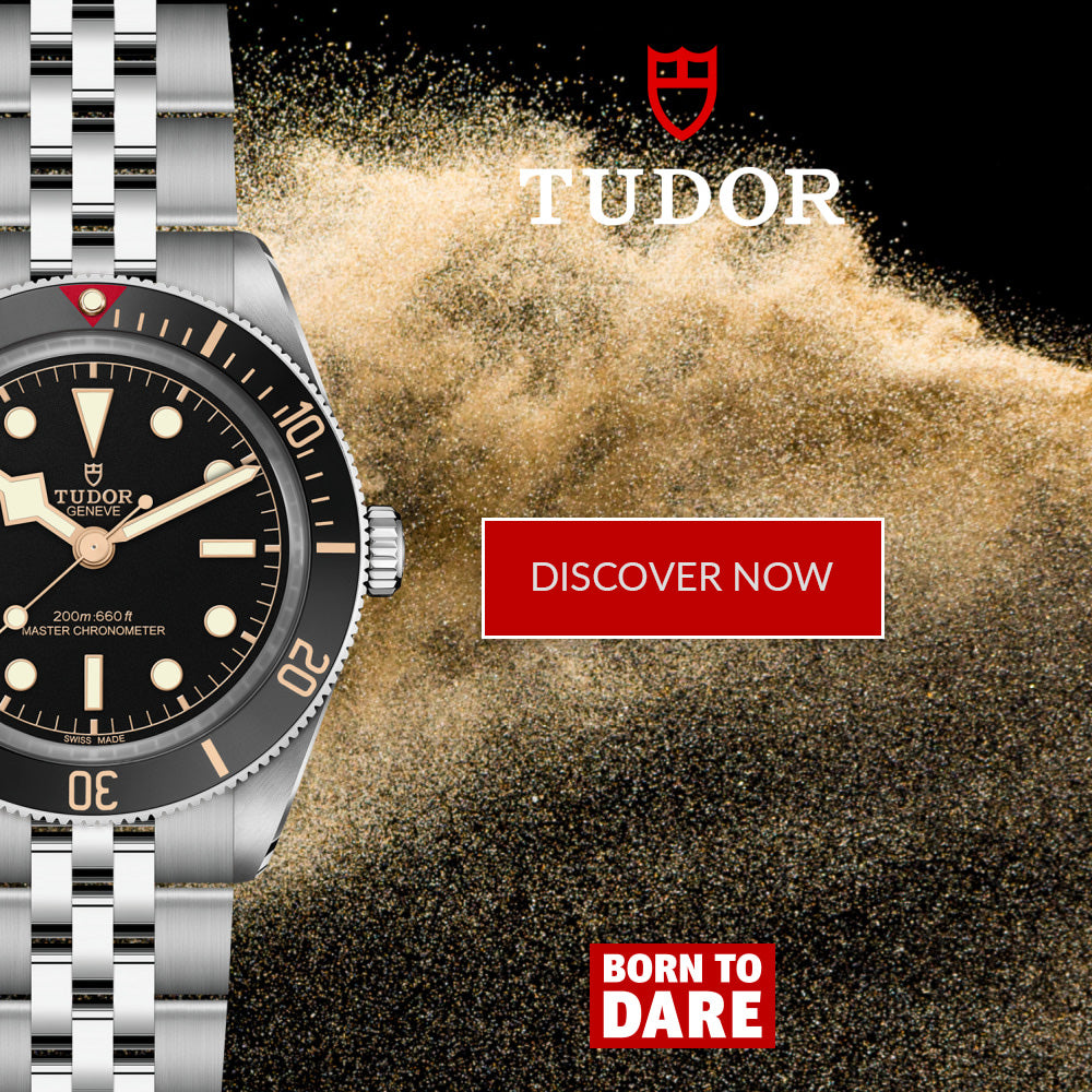 A Tudor Submariner Date watch with a silver stainless steel bracelet and black dial, shown alongside a dramatic background of sand and promotional text including 'TUDOR', 'DISCOVER NOW', and 'BORN TO DARE'.